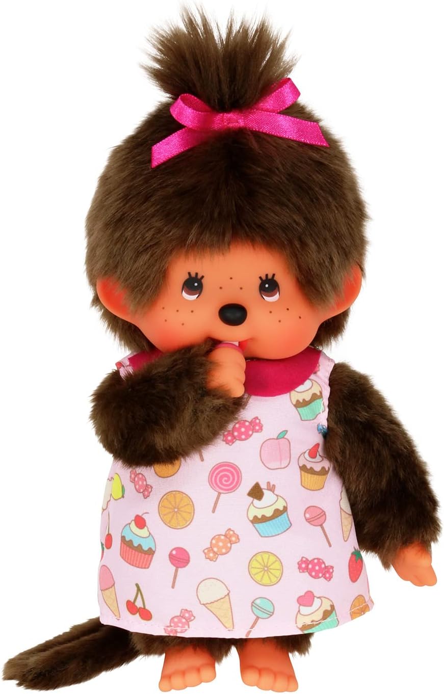 Peluche Monchhichi Fille Pop 'N Candy suçant son pouce, portant une robe imprimée gourmande et une couette attachée par un ruban rose.