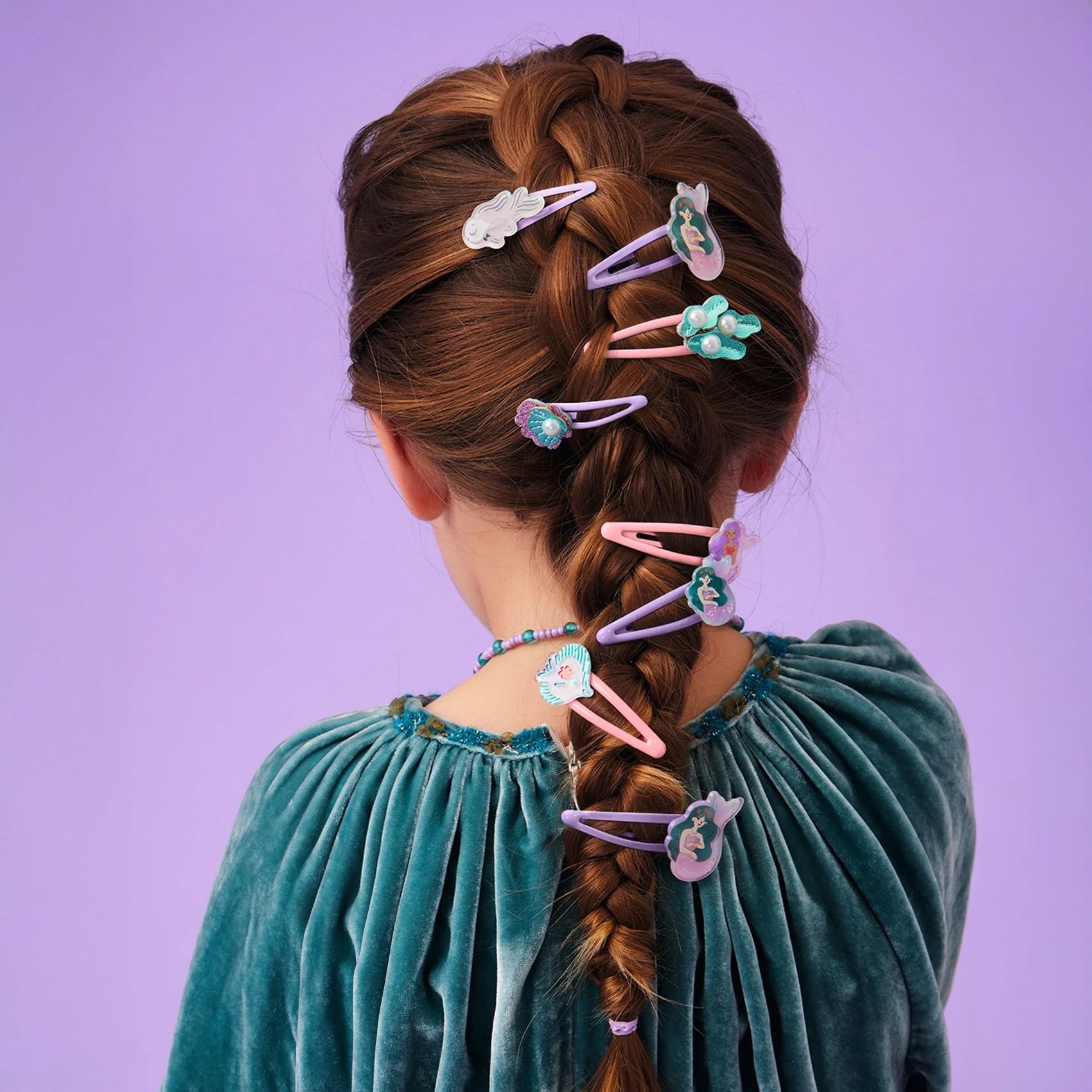 Jeune fille vue de dos avec une longue tresse française décorée de plusieurs barrettes à cheveux rose et violettes de la collection Plume par Djeco, sur le thème de la mer et des sirènes.