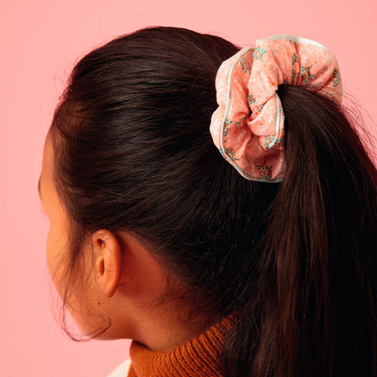 Jeune fille vue de profil et de dos portant une queue-de-cheval haute attachée avec un chouchou en tissu rose Pêche à motifs floraux et bordure vert d'eau, issu de la collection d'accessoires pour cheveux Plume de Djeco.