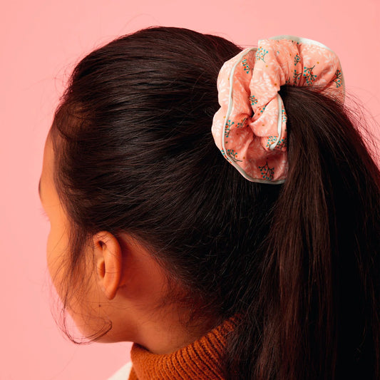 Jeune fille vue de profil et de dos portant une queue-de-cheval haute attachée avec un chouchou en tissu rose Pêche à motifs floraux et bordure vert d'eau, issu de la collection d'accessoires pour cheveux Plume de Djeco.