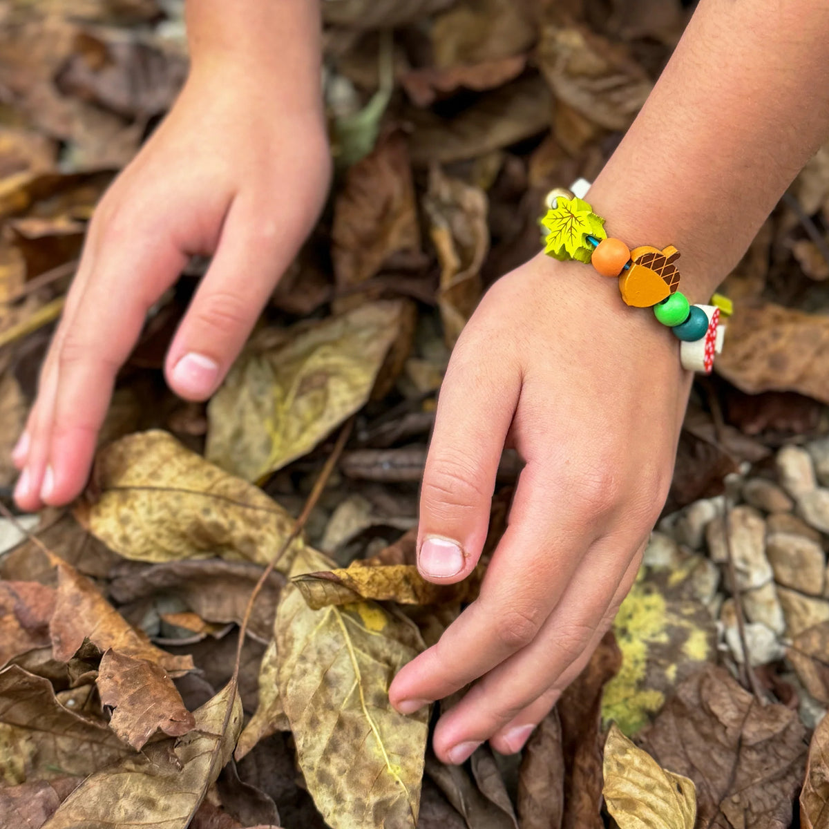 Poignet d'enfant portant le bracelet en perles de bois motifs forêt Cotton Twist au-dessus d'un lit de feuilles mortes d'automne.