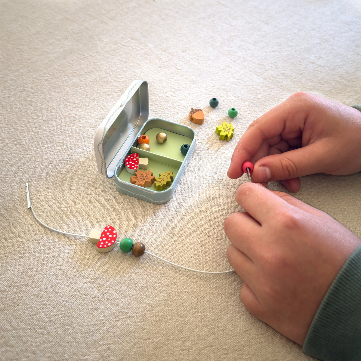 Mains d'un enfant en train d'enfiler des perles en bois colorées sur un cordon pour fabriquer un bracelet du kit Cotton Twist.