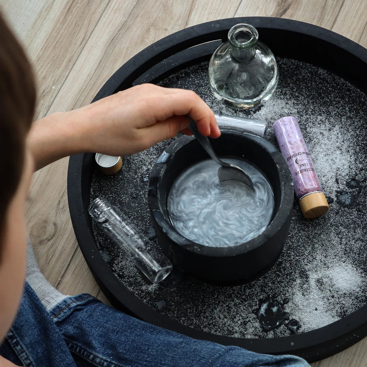Enfant mélangeant une potion magique nacrée et argentée dans un mortier noir avec une cuillère, entouré de fioles d'ingrédients Les Petits Sorciers.