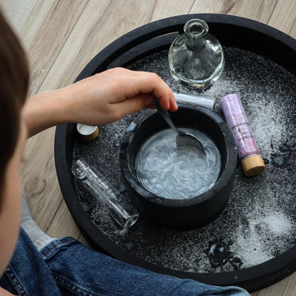Enfant mélangeant une potion magique nacrée et argentée dans un mortier noir avec une cuillère, entouré de fioles d'ingrédients Les Petits Sorciers.