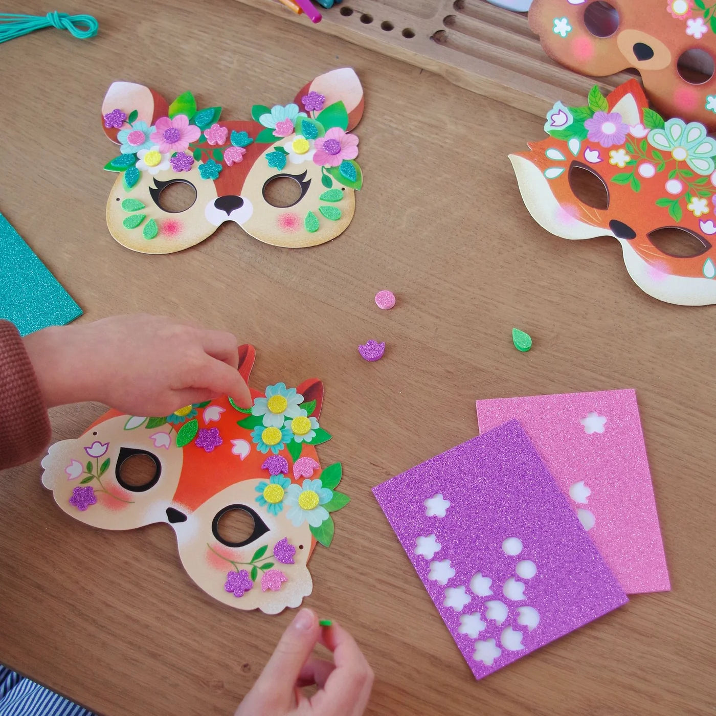 Enfant décorant des masques d'animaux de la forêt avec des mosaïques autocollantes pailletées de la collection Do It Yourself de Djeco.