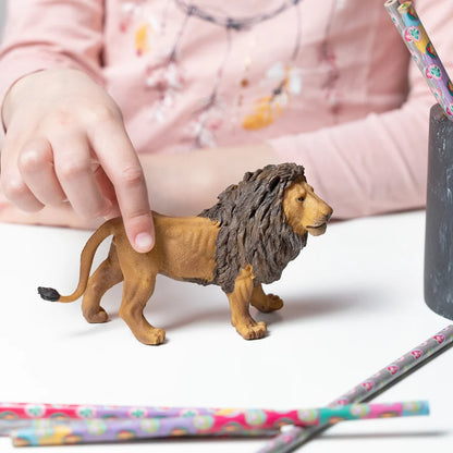 Figurine de lion majesteux Papo peinte à la main posée sur une table et tenue par la main d'un enfant.