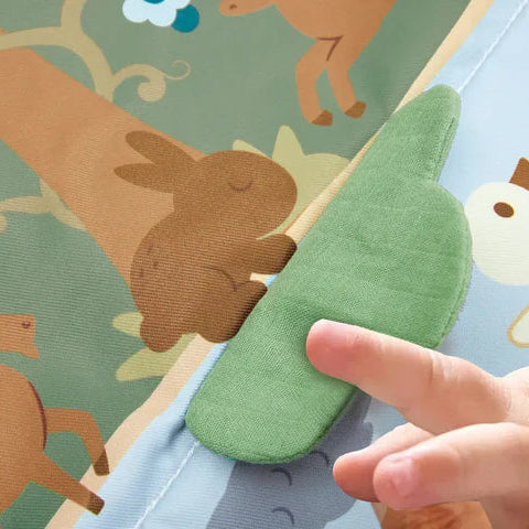 Gros plan sur la main d'un bébé touchant un élément texturé en tissu vert en forme de buisson sur le tapis d'éveil Hape.