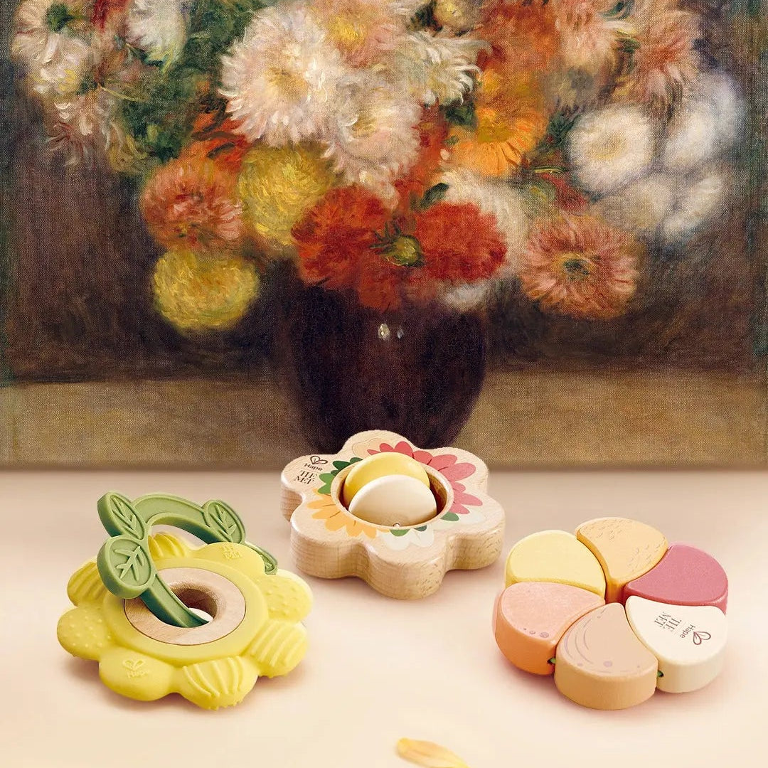 Ensemble de trois hochets en bois et silicone en forme de fleurs posés devant une reproduction d'un tableau de bouquet de fleurs de Renoir.
