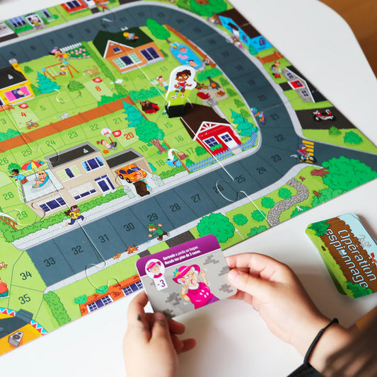 Une personne tient une carte de jeu spéciale du jeu Opération espionnage de Placote au-dessus d'un plateau de jeu illustrant un quartier résidentiel.