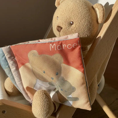 Livre en tissu pour bébé Marcel l'Ours Egmont Toys tenu par un ourson en peluche sur une chaise en bois.