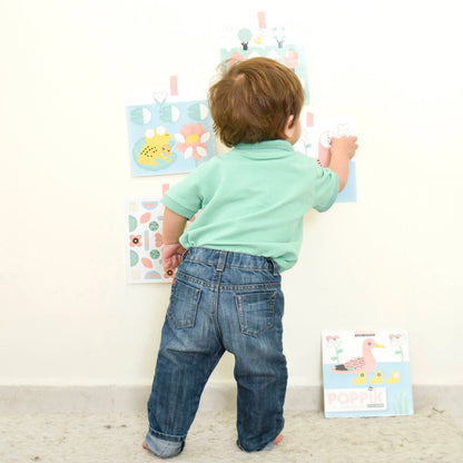 Un jeune enfant de dos colle les fiches illustrées de l'ensemble d'autocollants « Ma première rivière » de Poppik sur un mur blanc, montrant le jeu en cours d'utilisation.