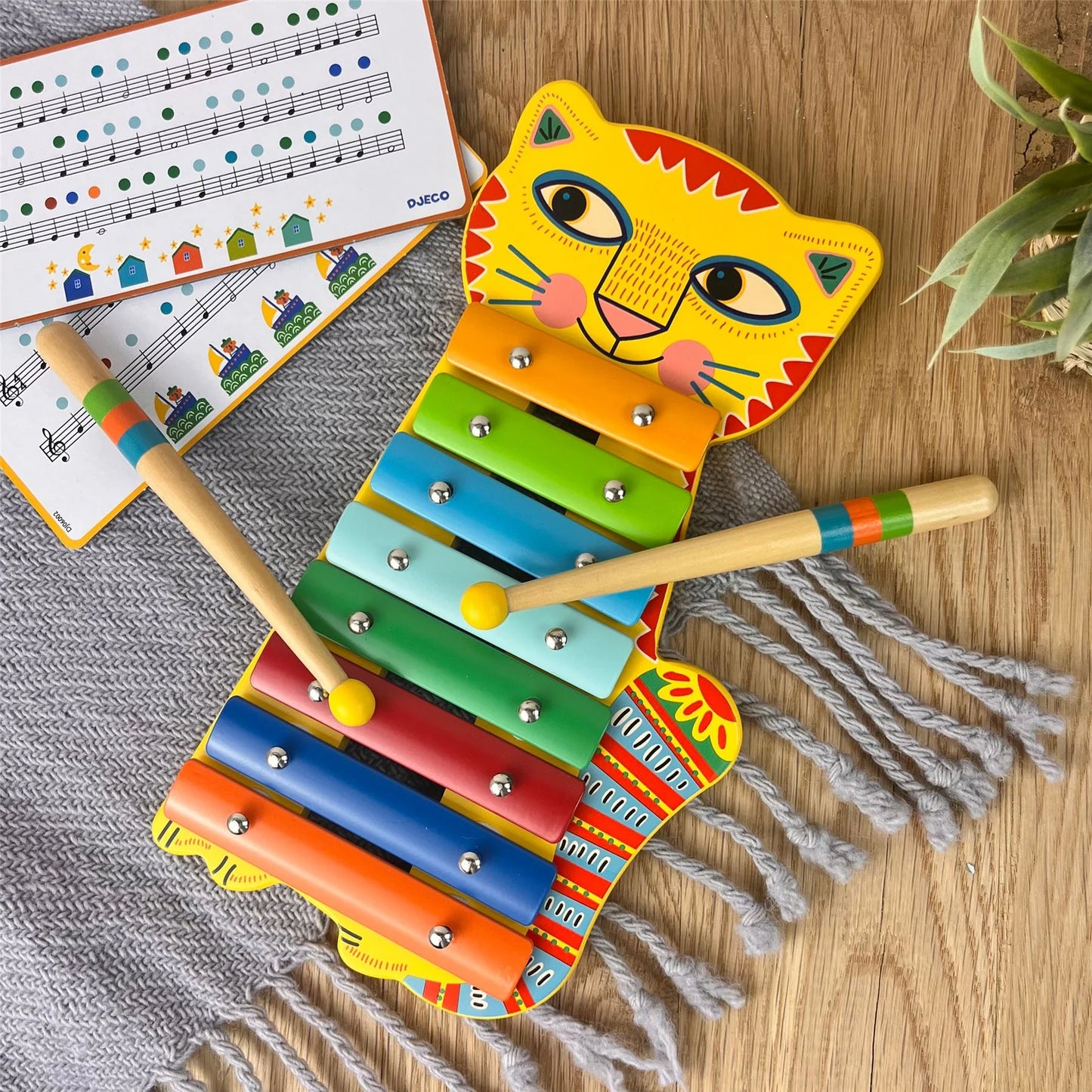 Métallophone en bois coloré en forme de chat jaune de la gamme Animambo par Djeco, posé à côté de ses baguettes et de partitions musicales simplifiées.