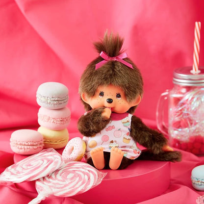 Peluche Monchhichi fille Pop 'N Candy de Sekiguchi assise dans un décor gourmand rose composé de macarons, sucettes et friandises.