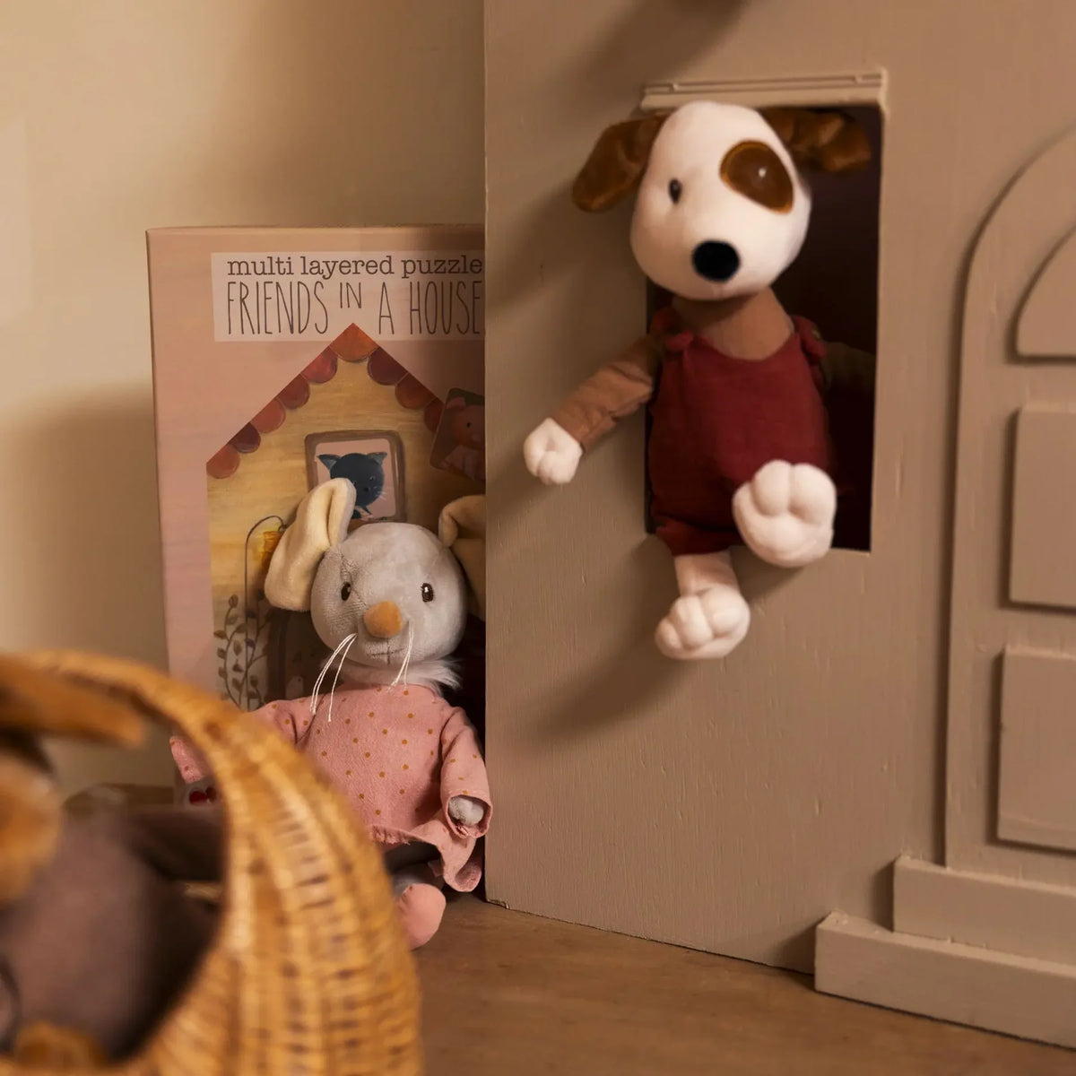 La peluche chien Eddie qui regarde par la fenêtre d'une maisonnette en bois, à côté de la souris Agathe et d'un casse-tête de la marque.