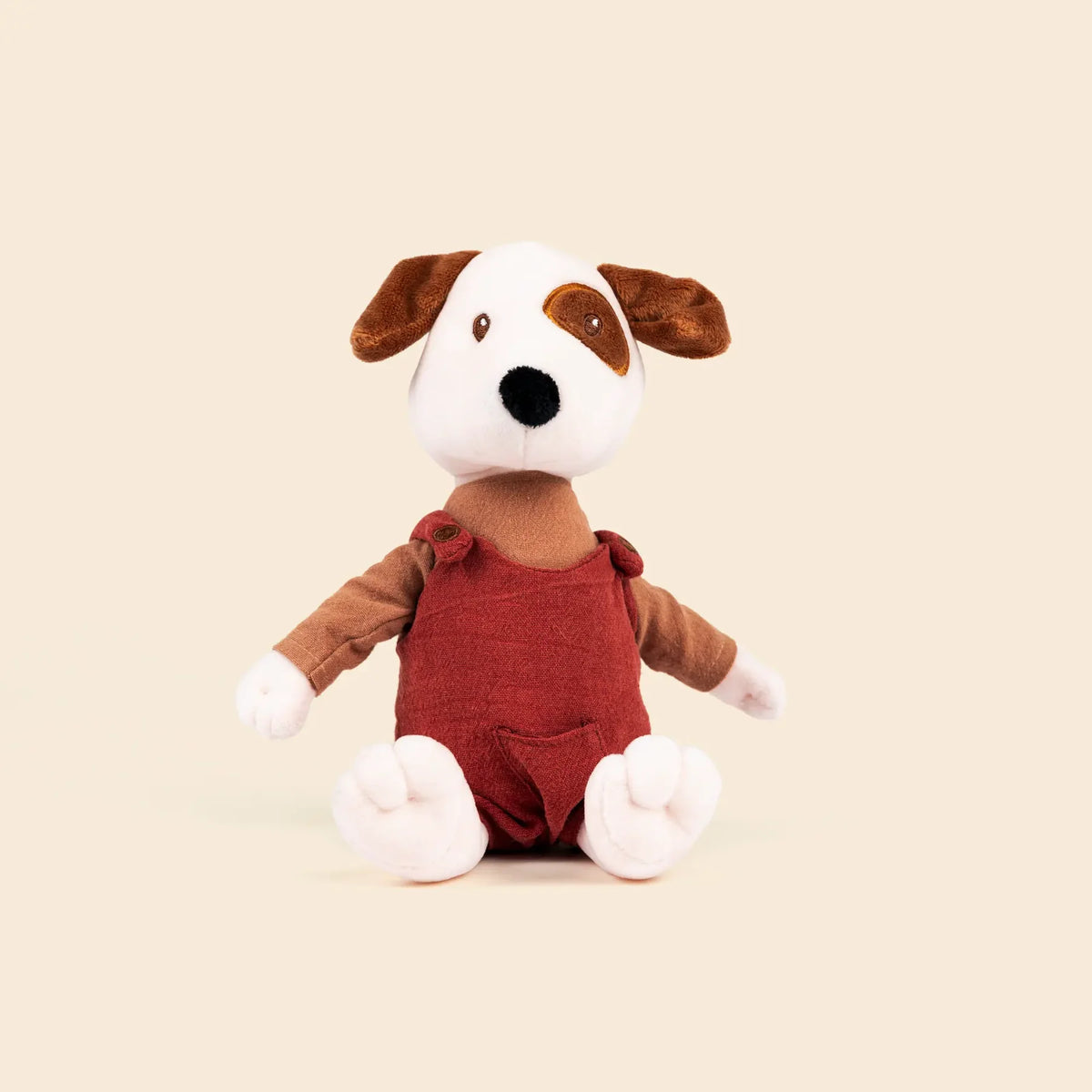 Peluche Eddie le chien de Egmont Toys, assis, avec une tache brune sur l'œil et vêtu d'une salopette rouge brique en gaze de coton.