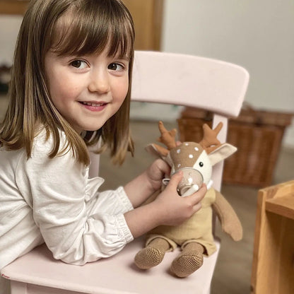 Une petite fille souriante assise sur une chaise rose, tenant la peluche faon Woody dans ses mains.