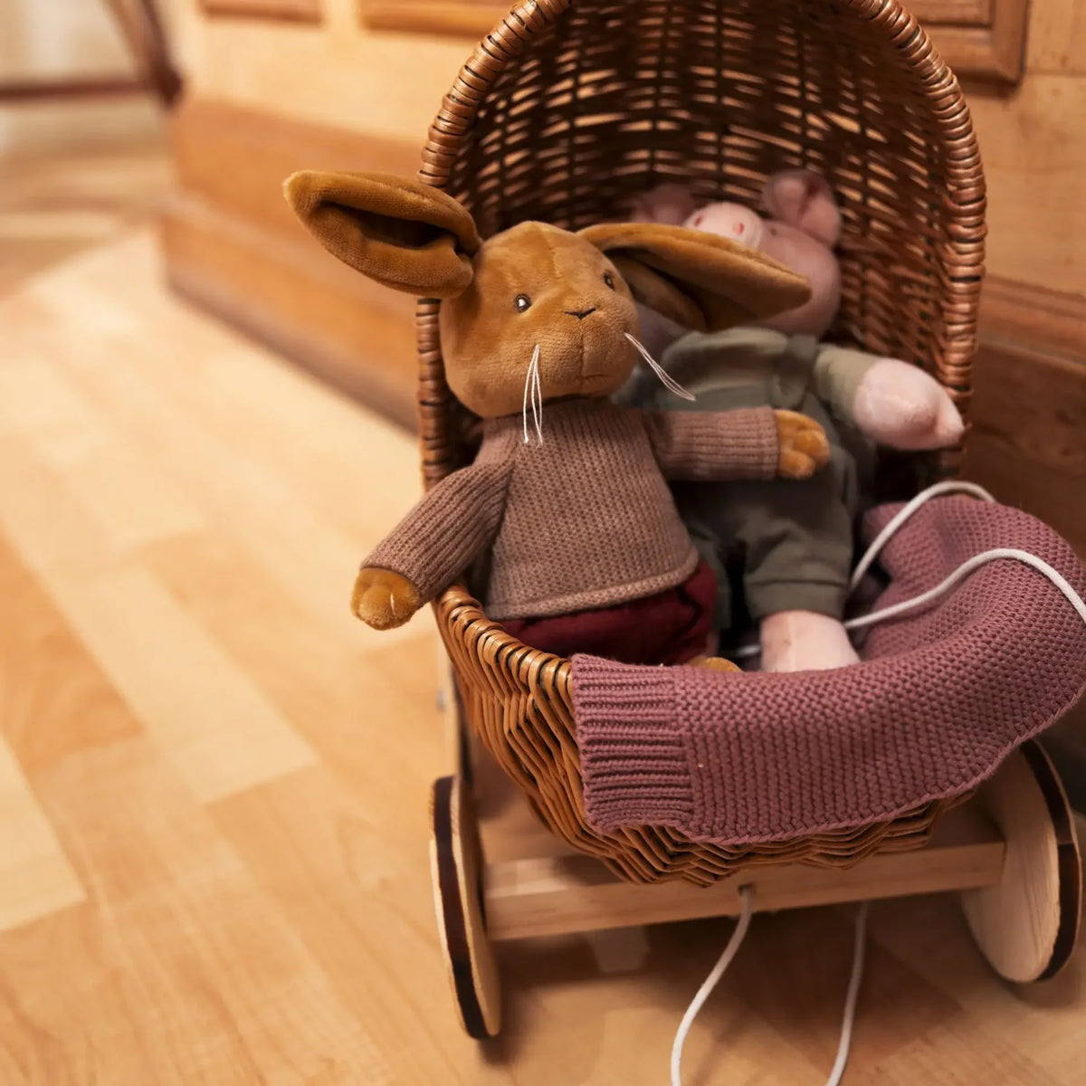 La peluche lapin Lenny installée dans un petit landau en osier aux côtés d'une peluche cochon, avec une couverture en tricot assortie.