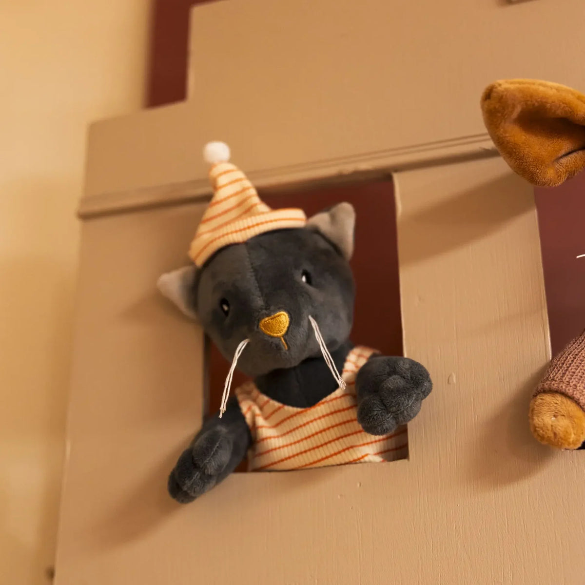 Peluche Malo le chat gris Egmont Toys passant la tête par la fenêtre d'une cabane en bois.