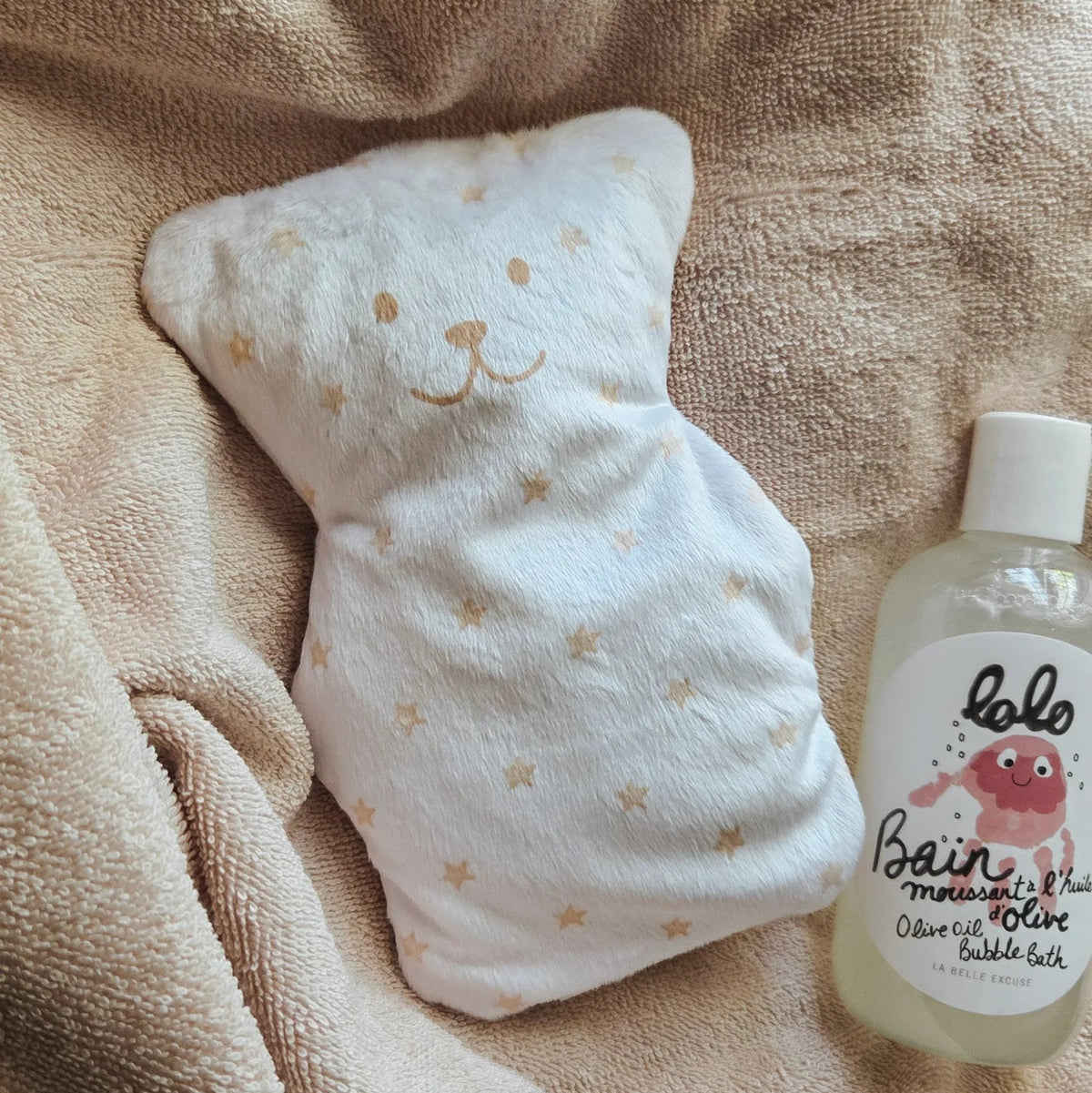 Petit ourson thérapeutique Béké Bobo à motif d'étoiles posé sur un plaid moelleux à côté d'une bouteille de produit pour le bain Lolo.