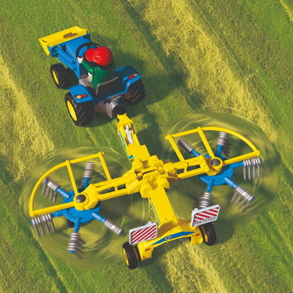 Vue du dessus du tracteur bleu Playmobil tirant l'andaineur à foin jaune aux toupies rotatives dans un champ d'herbe.