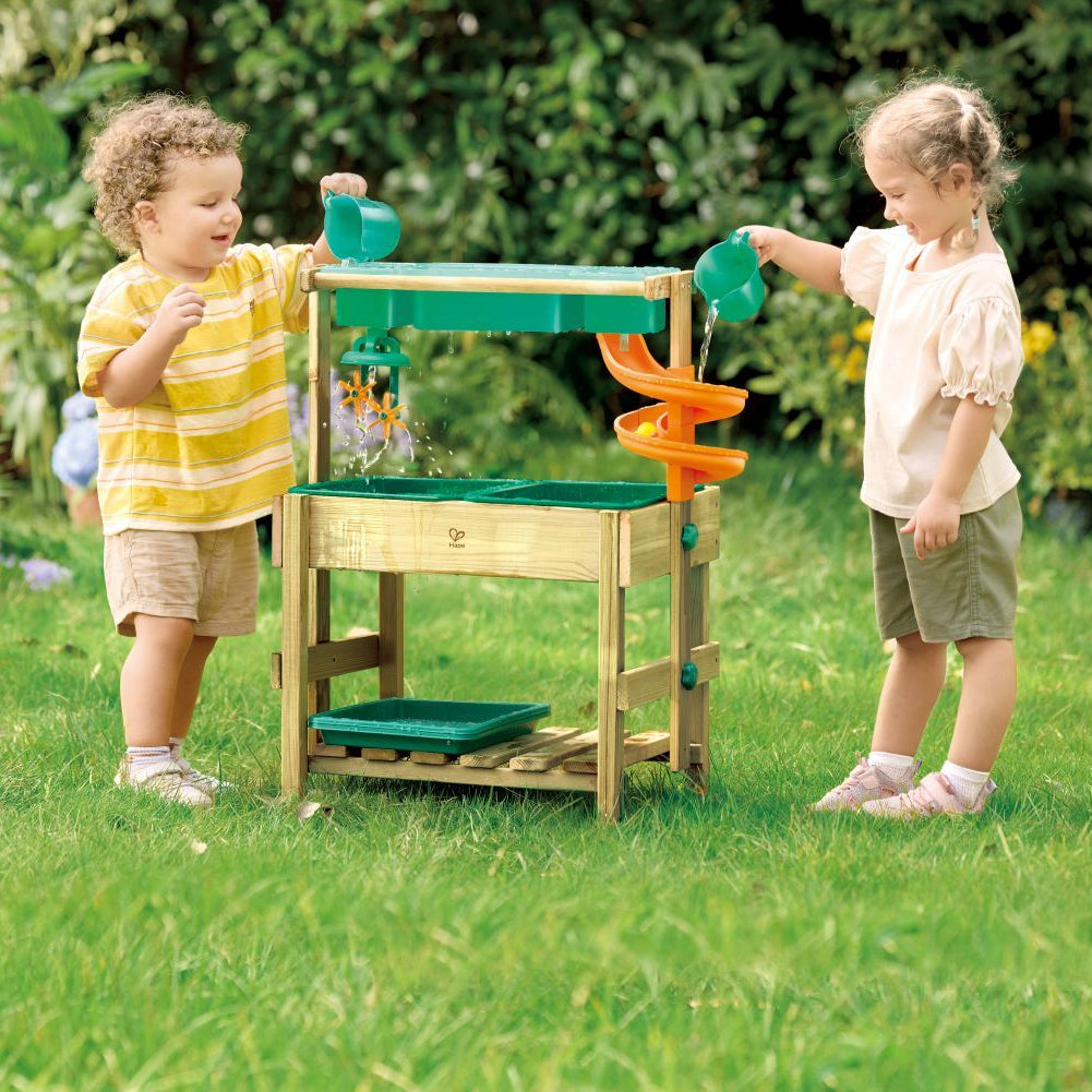 Deux jeunes enfants s'amusent dehors dans le jardin avec la table de jeu d'eau en bois Hape, versant de l'eau à l'aide de pichets pour activer les engrenages et le toboggan.