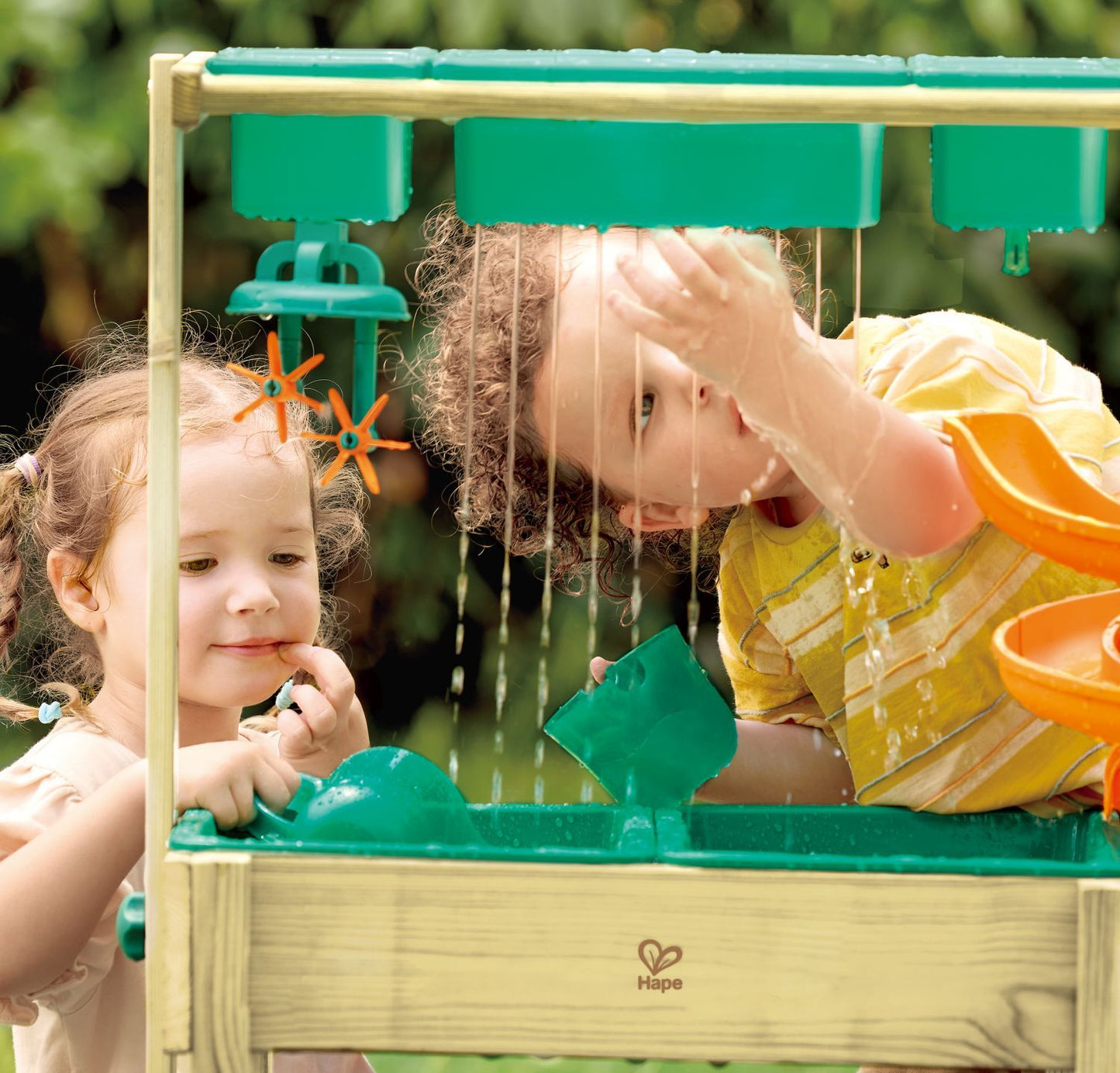 Gros plan sur deux enfants jouant sous l'effet de pluie fine de la table d'eau extérieure Hape, observant l'eau couler à travers le bac suspendu.