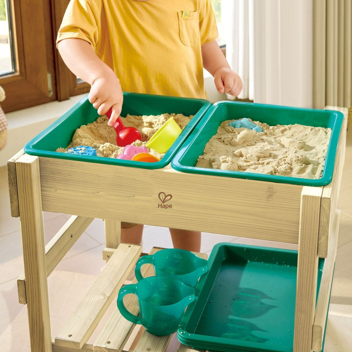Un petit garçon utilise la table en bois Hape comme table sensorielle à l'intérieur de la maison, remplie de sable de jeu avec des moules et des pelles colorées.