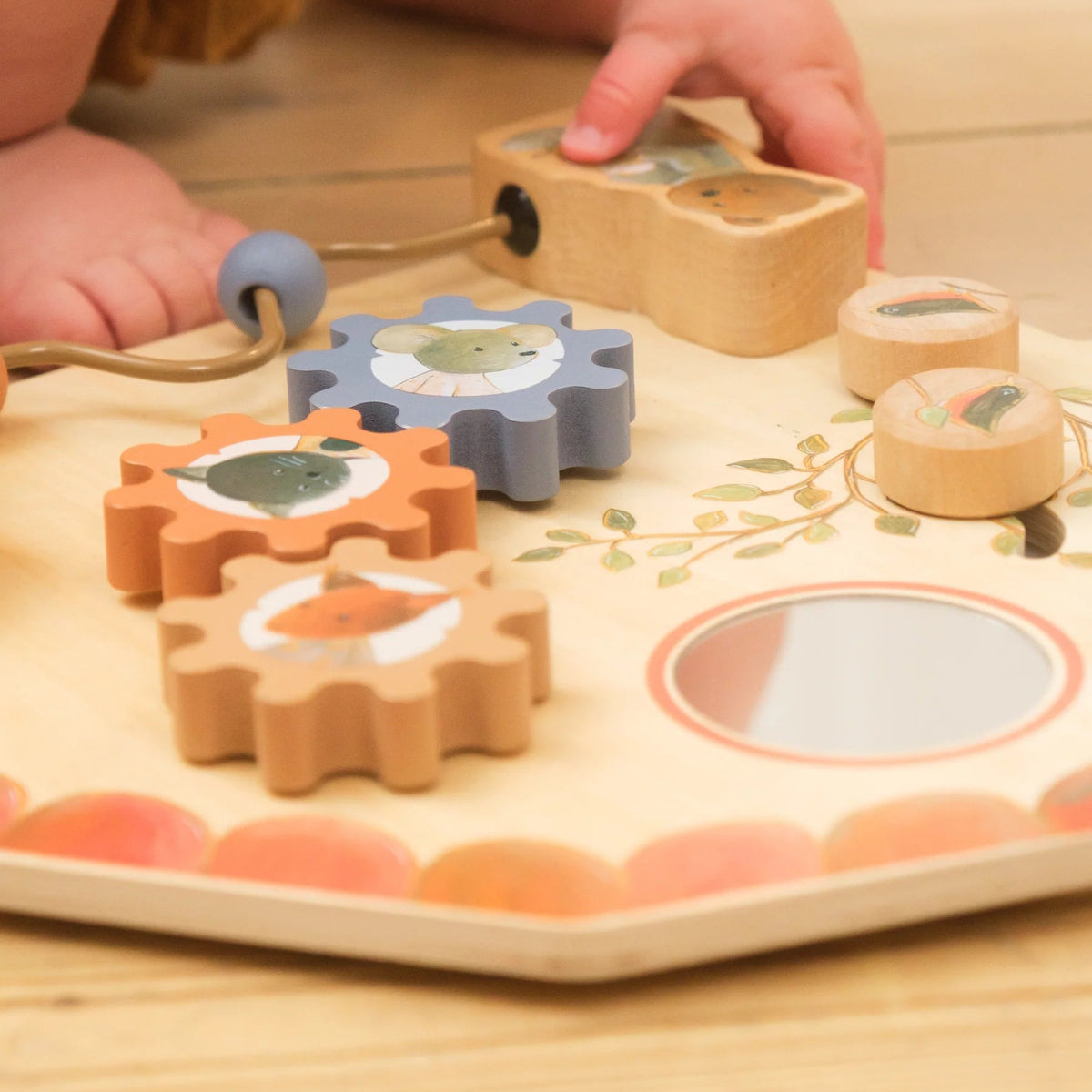 Mains d'un bébé manipulant un tableau d'activités en bois de Egmont Toys posé au sol, montrant des engrenages colorés avec des illustrations d'animaux.