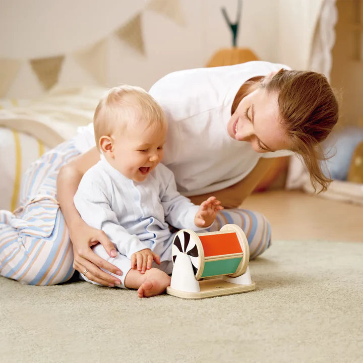 Un bébé assis par terre avec sa maman qui rit en faisant tourner le tambour d'éveil en bois Hape installé horizontalement sur son support.
